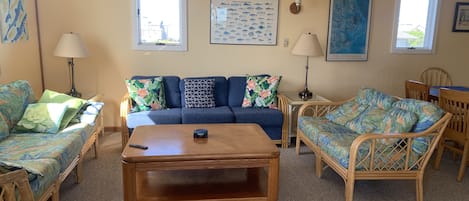 A living room with a blue sofa, a wooden coffee table, and wicker furniture. There are two lamps, a framed fish poster, and a painting on the wall.