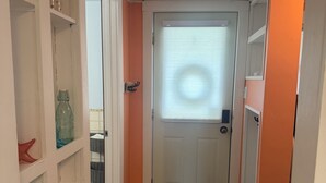 Un pasillo con una puerta blanca, una ventana con persianas, suelo de madera y un tapete con motivos de cangrejos.