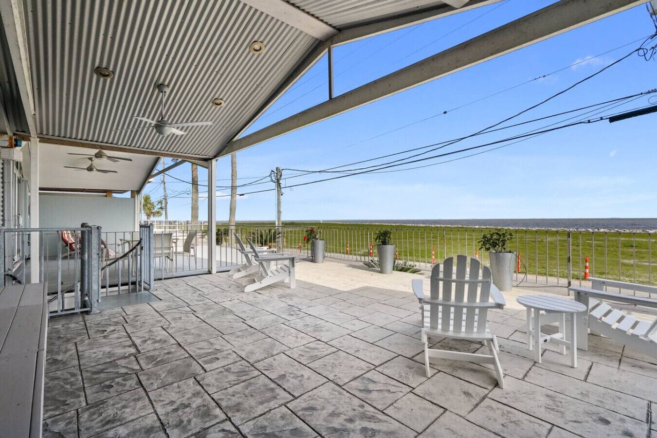 A covered outdoor area with a view of the beach, featuring a patio with chairs and a table, a ceiling fan, and a railing.