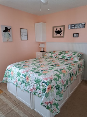 Une chambre avec une couvre-lit à motifs floraux, une armoire blanche, une table de chevet avec une lampe et des œuvres d’art murales, notamment un oiseau et un tableau encadré avec du texte.