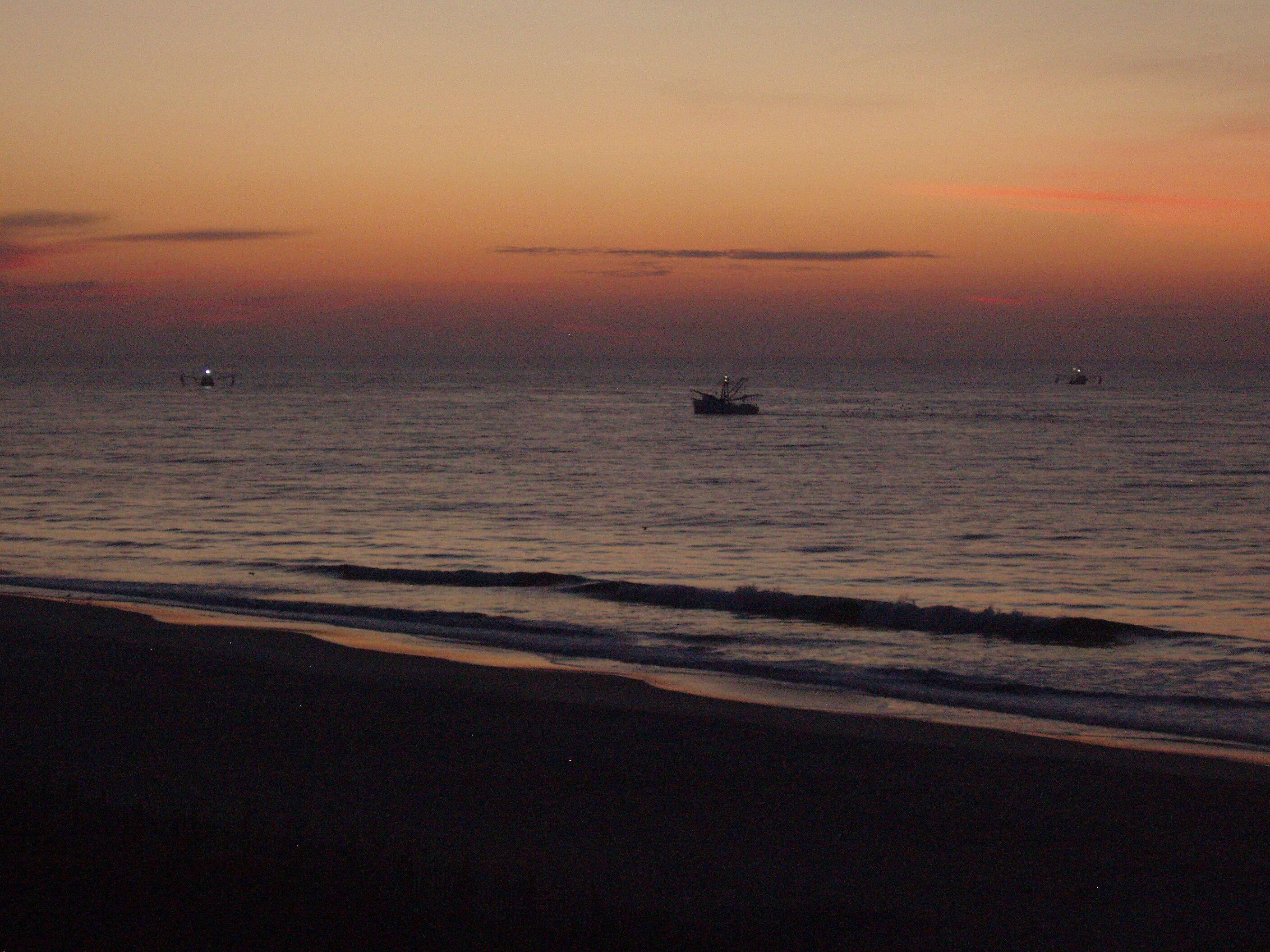 Una platja al capvespre amb aigües tranquil·les i alguns vaixells.