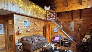Ein gemütliches Blockhaus-Interieur mit Holzboden, einem braunen Ledersofa, einem Kuhfellteppich und einer Treppe, die zu einer oberen Ebene führt.