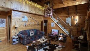 Ein gemütliches Blockhaushaus-Interieur mit Holzbalkendecke, Deckenventilator, Treppe, einer Couch, einem Couchtisch und einem Kuhfellteppich.