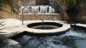Un bain à remous avec un mur de pierres en arrière-plan, un coin salon avec des coussins à rayures et une plante en pot.