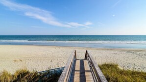En träpromenad som leder till en sandstrand med havet i bakgrunden.