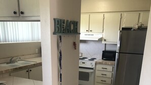 Une cuisine avec un plan de travail en bois, un évier, une cuisinière et un réfrigérateur. On y trouve deux tabourets et une enseigne fixée au mur sur laquelle est écrit « PLAGE ».