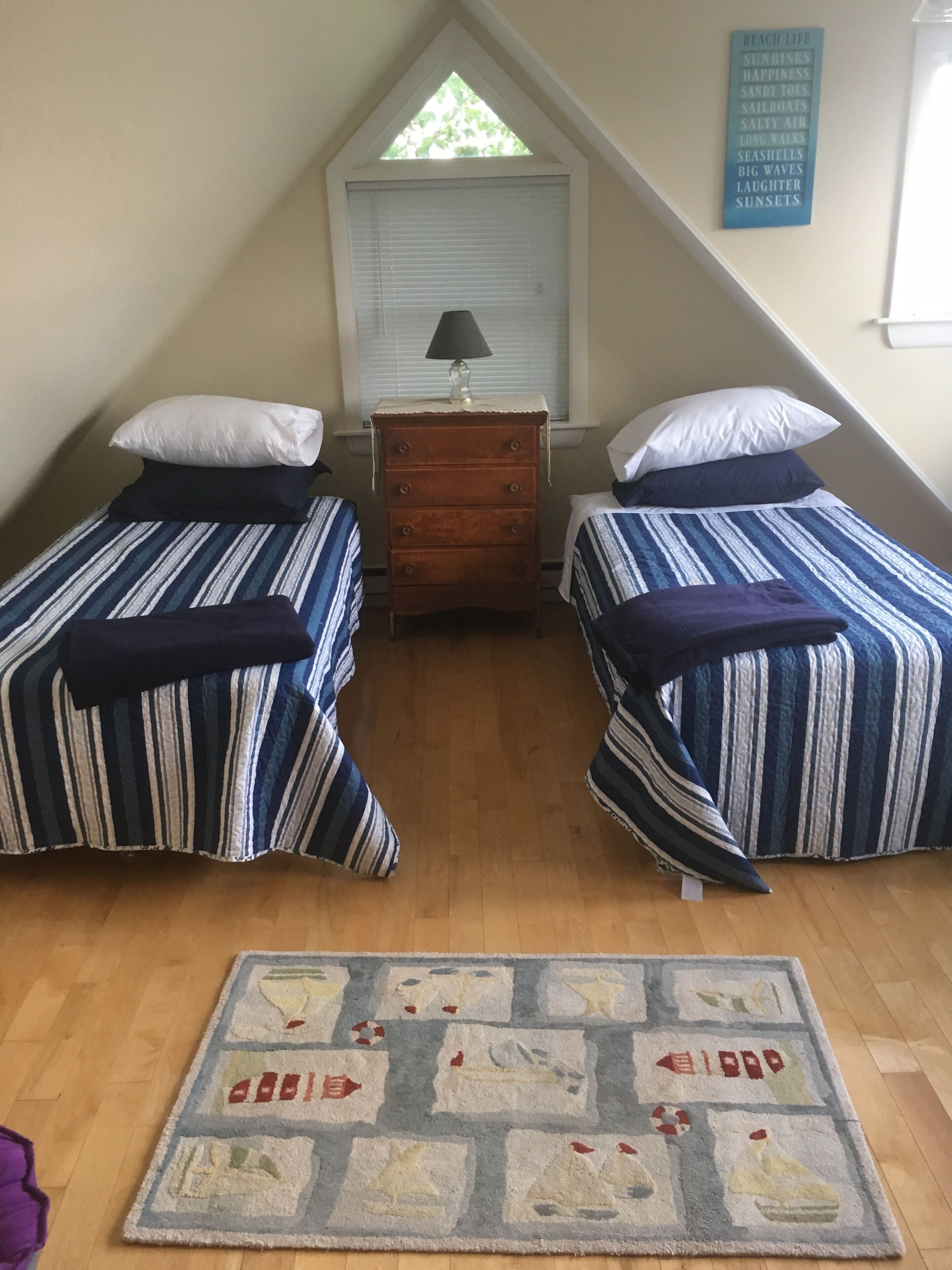 Een knus, klein kamertje met twee bedden, een houten dressoir, een raam met zonwering en een vloerkleed met strandthema.