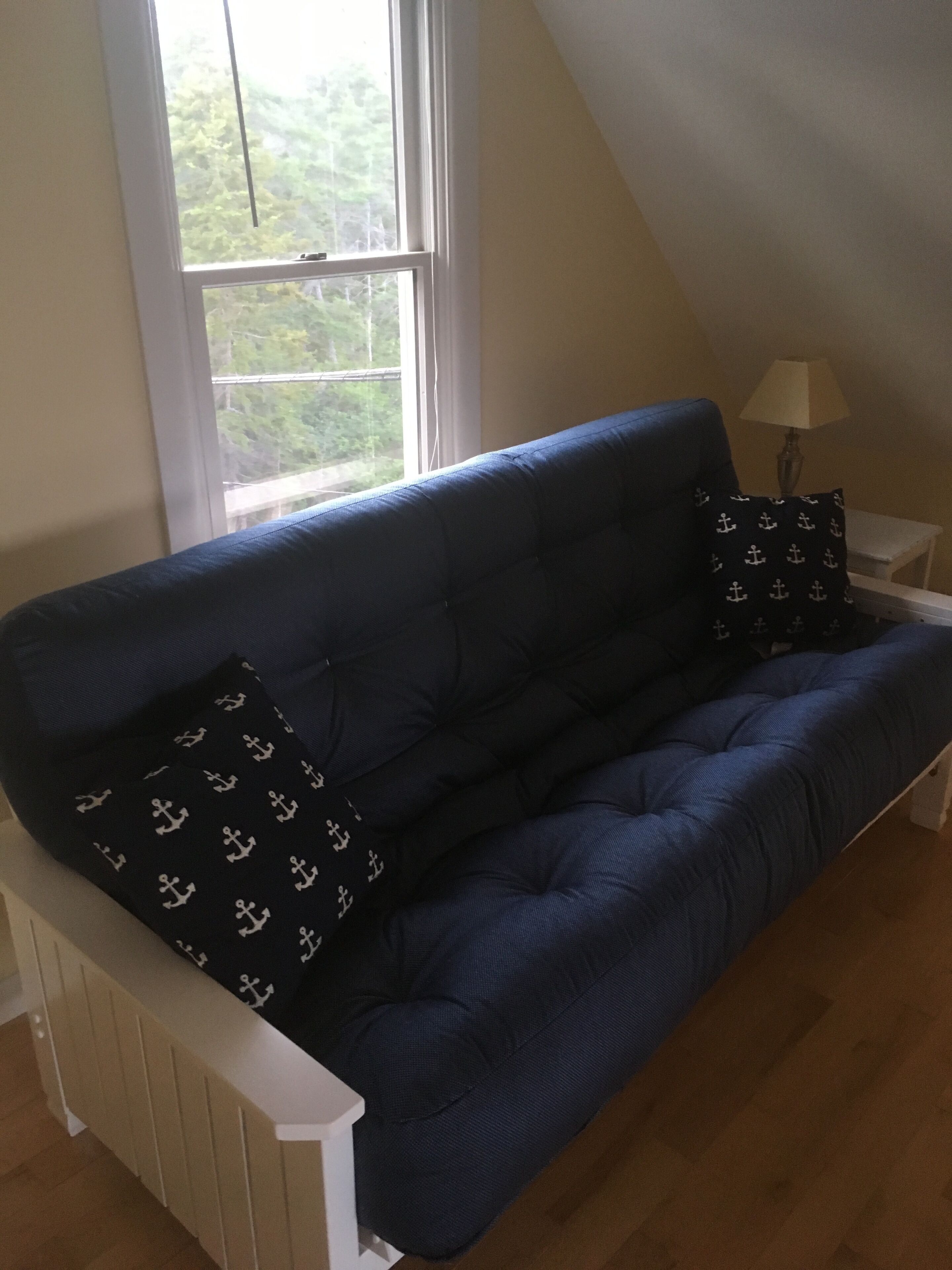 A blue sofa with patterned pillows, a white side table with a lamp, and a window with a view of greenery.