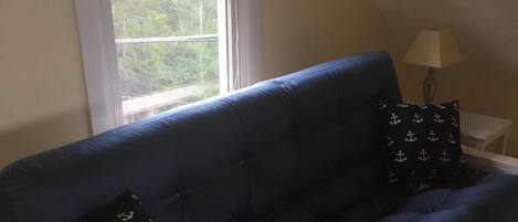 A blue sofa with patterned pillows, a white side table with a lamp, and a window with a view of greenery.