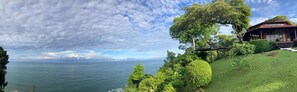 A coastal landscape with a house overlooking the sea, surrounded by lush greenery and a clear sky.