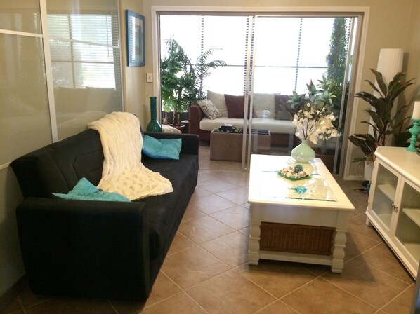 A living room with a black sofa, a white coffee table, and a sliding glass door leading to a balcony with plants.