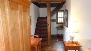 Una escalera de madera que sube a un segundo piso, una silla de madera, una pequeña mesa con una lámpara y un radiador en la pared.