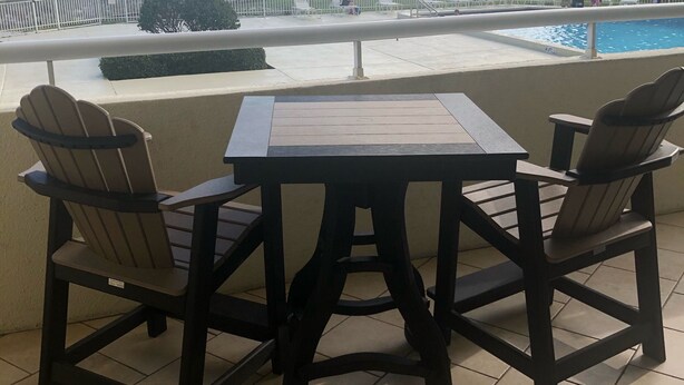 A balcony with two chairs and a table, overlooking a swimming pool and beach.
