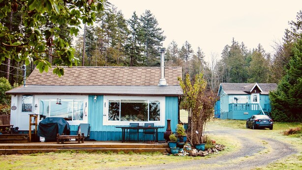 A small, single-story building with a blue exterior, a covered patio area with a table and chairs, and a car parked in the driveway.