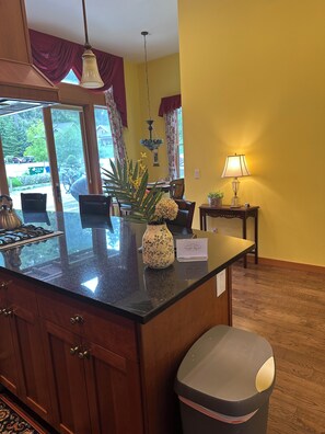 A kitchen with wooden cabinets, a granite countertop, a vase with flowers, a hanging light fixture, and a view of the outdoors through large windows. - Captain's Suite - 2 Bedroom Condo in Eastsound across from the park (Eastsound)
