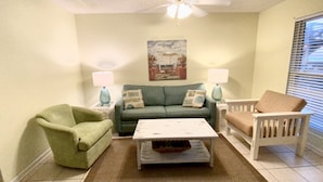 Un salón con un sofá verde, una mesa de centro blanca y un sillón verde. Hay dos lámparas sobre mesitas auxiliares, un cuadro en la pared y un ventilador de techo.