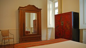 Ein Schlafzimmer mit einem Holzschrank, einem Spiegel, einem Korbstuhl und einem Bett mit gestreifter Bettdecke.