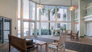 A spacious lobby with large windows, wooden furniture, and a view of palm trees outside. - Regency Towers 707 East Tower   Fall is Better at the Beach!  (Pensacola Beach)