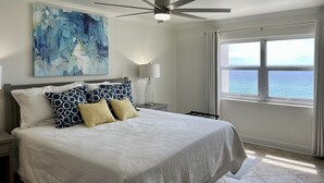 A bedroom with a bed, a ceiling fan, a nightstand, a lamp, a window with a view of the ocean, and an abstract painting on the wall. - Regency Towers 707 East Tower   Fall is Better at the Beach!  (Pensacola Beach)