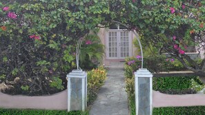 A stone pathway leading to a building with a white door, surrounded by lush greenery and flowering plants. - The Perfect Home to Enjoying St. Lucia (Rodney Bay)