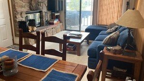 A cozy living room with a stone fireplace, a flat-screen TV, a blue sofa, and a dining area with wooden furniture. - Great family ski condo with access to all the Fraser valley has to offer (Fraser)