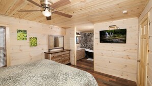 Interior de una cabaña de madera con cama, ventilador de techo, baño con pared de piedra y televisor en la pared.