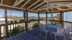 A covered outdoor area with a wooden deck, white metal chairs, and a round table.