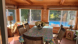 A wooden dining area with a table covered in a colorful tablecloth, surrounded by wicker chairs, a vase with flowers, and a view of a swimming pool through large windows. - Key West in Cape May: Hot tub open year round! Dog friendly! (Villas)
