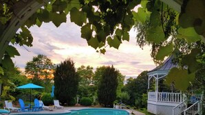 A swimming pool with lounge chairs and umbrellas, surrounded by greenery and a gazebo.