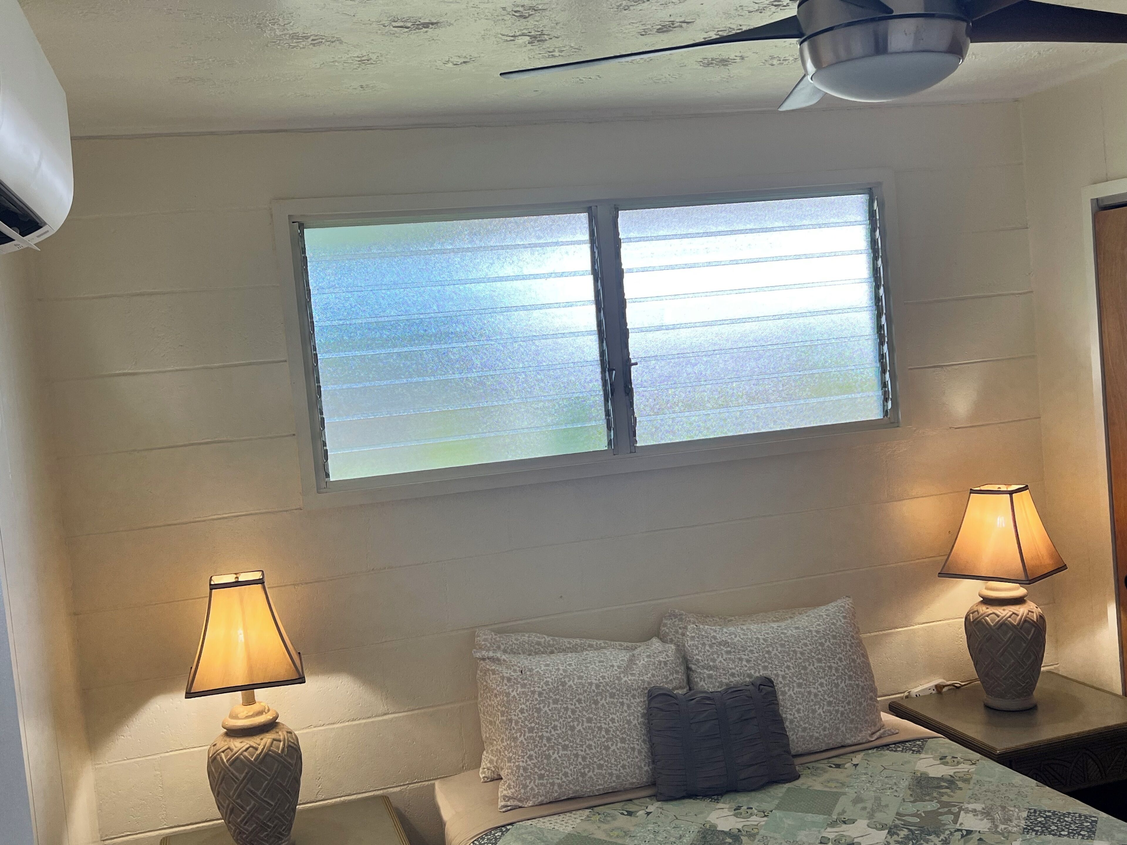 A room with a bed, two bedside lamps, a window with blinds, a ceiling fan, and an air conditioning unit.