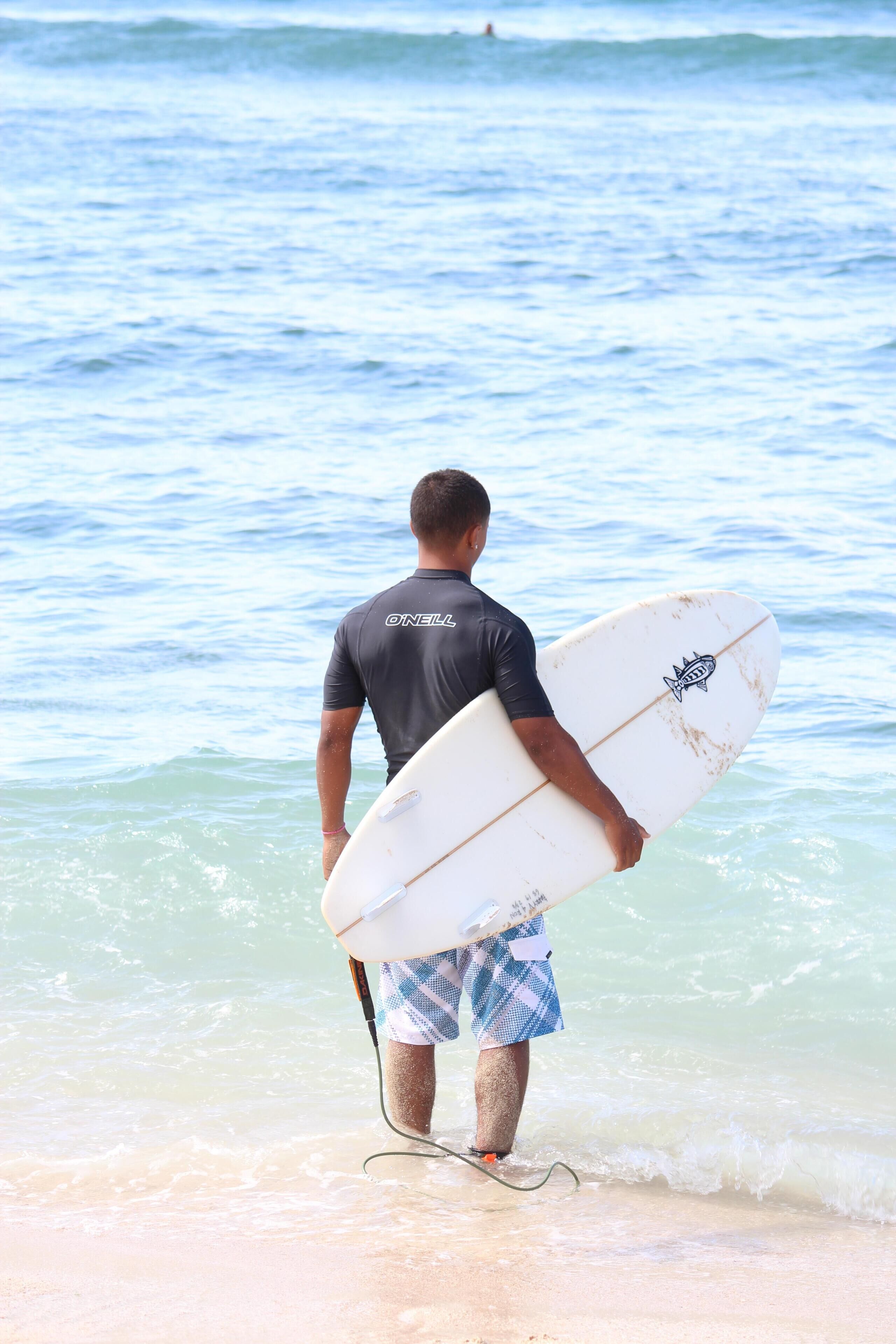 A man carrying a surfboard, wearing a dark wetsuit and blue shorts, walking towards the ocean.