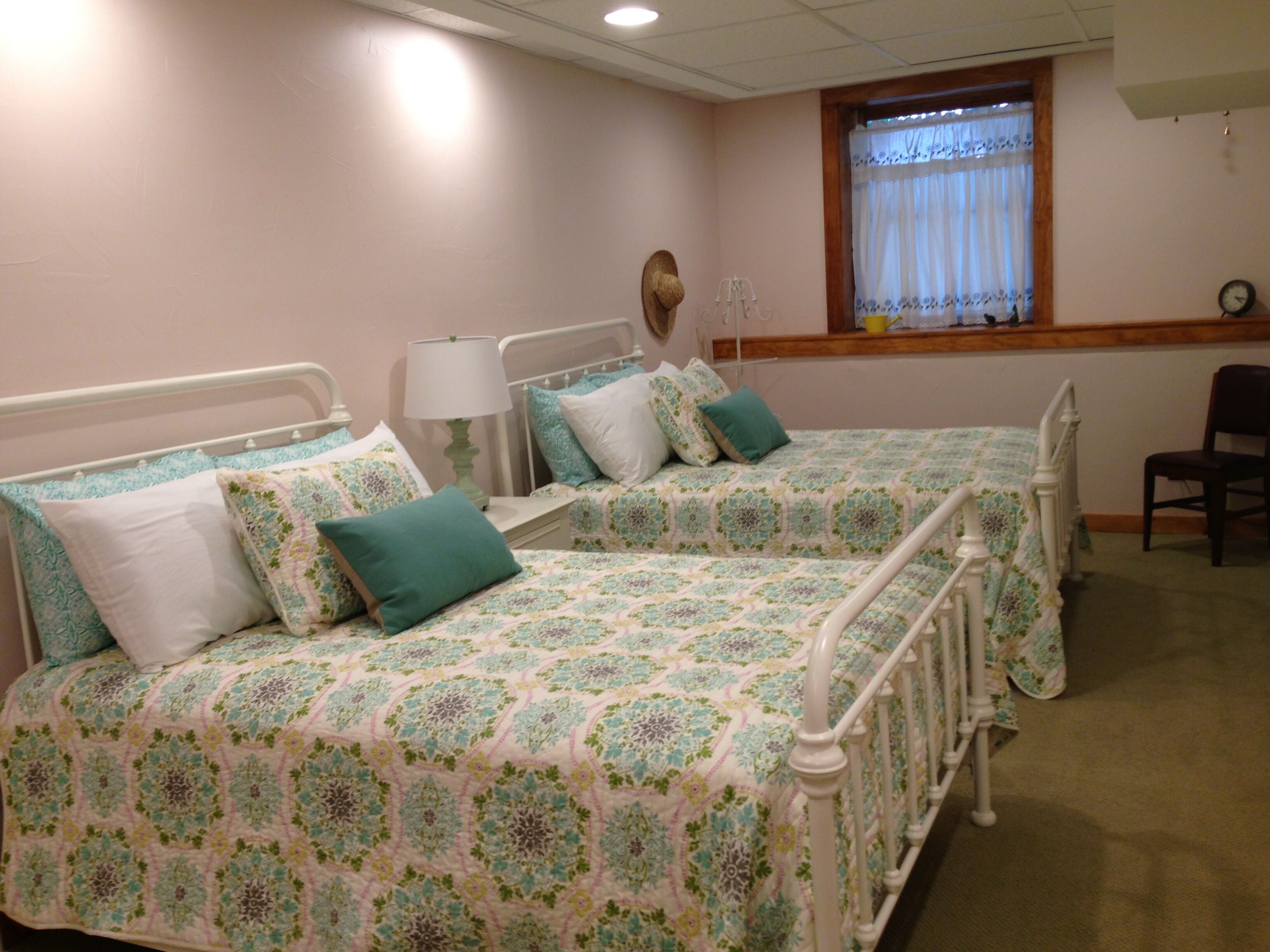 Ein Zimmer mit zwei Betten, jeweils mit gemusterten Bettdecken und Kissen, einem Nachttisch mit Lampe, einem Sessel und einem Fenster mit Vorhängen.