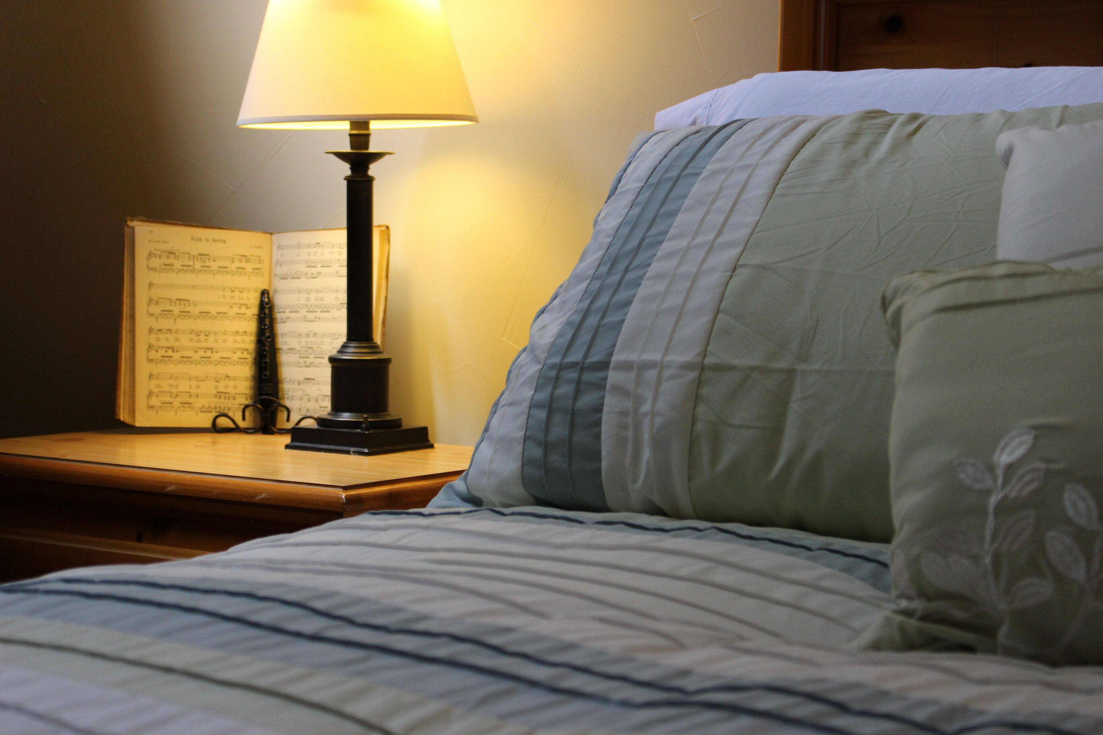 A neatly made bed with a striped comforter and pillows, a bedside table with a lamp and an open book of sheet music.