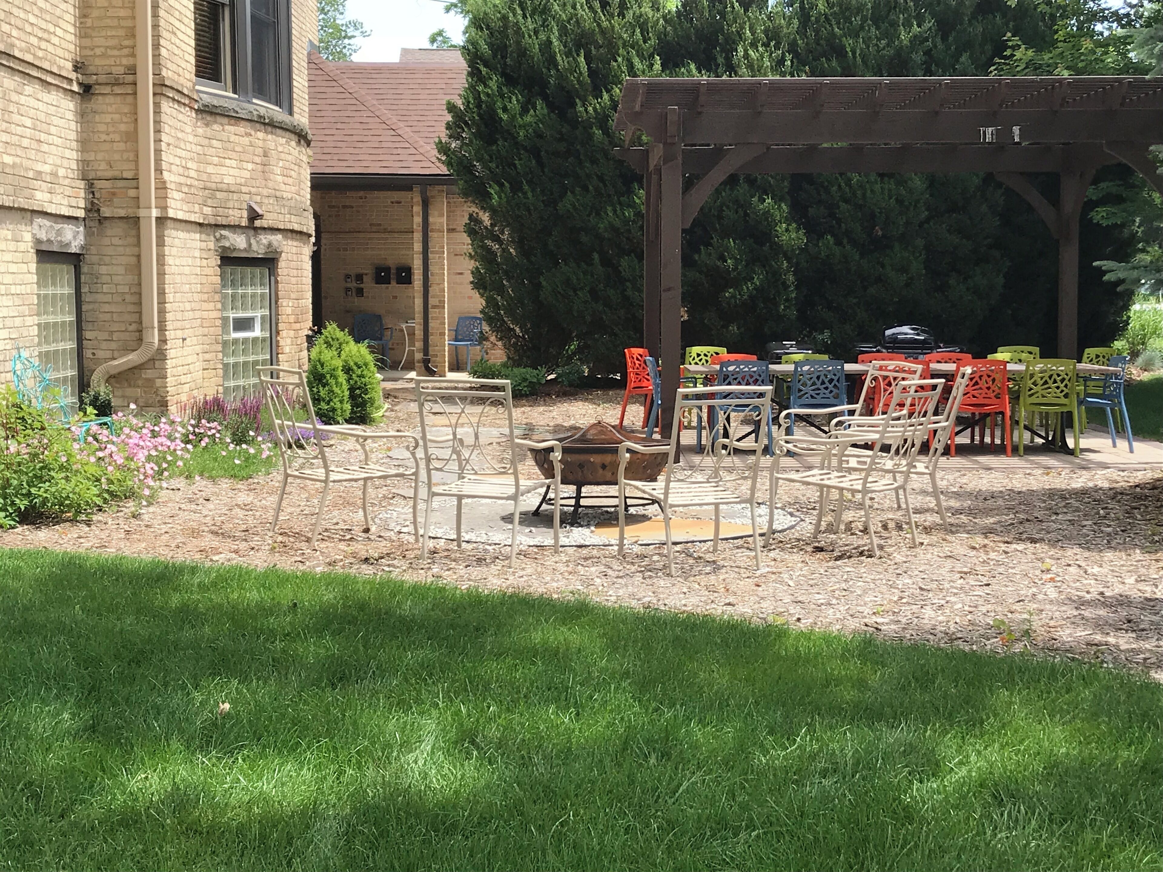 Un espace extérieur aménagé en terrasse, avec des chaises et des tables en métal, une pergola en bois et, au fond, un bâtiment en briques.