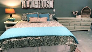 A bedroom with a bed, a nightstand with a lamp, a dresser, and a wall sign that reads "BEACHHOUSE".