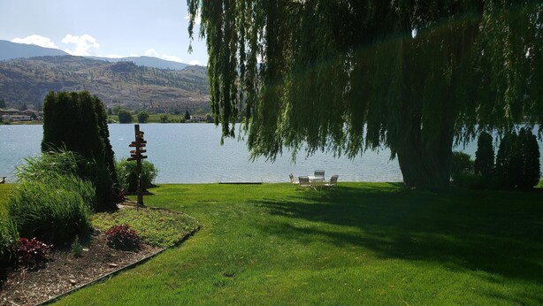 Un jardín tranquilo junto al lago, con un césped cuidado, una pequeña mesa y sillas bajo un sauce llorón, y una vista distante de colinas y montañas.