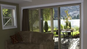 Una sala de estar con un sofá color beige, un muro de vidrio corredero que da a un patio con una mesa y sillas, y vistas a un lago y montañas.