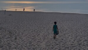 Une plage de sable où un enfant marche vers l’eau, quelques personnes visibles au loin et un coucher de soleil en arrière-plan.