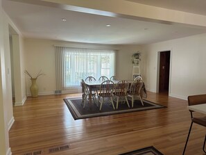 Un vaste espace dédié à la salle à manger, doté d’un plancher en bois, d’une table à manger avec des chaises et d’une grande fenêtre équipée de stores.