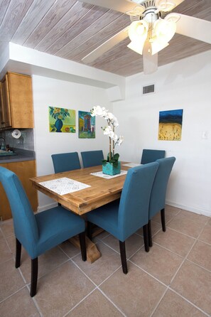 Uma área de refeições com mesa de madeira, cadeiras azuis e um ventilador de teto.