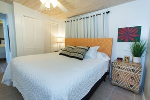 Quarto com uma cama, mesa de cabeceira, relógio na parede, planta em vaso e porta para outro cômodo.