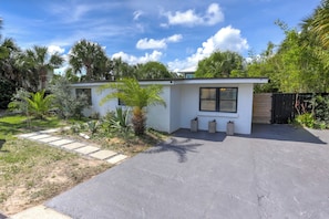 Uma casa moderna com calçada pavimentada, um pequeno jardim com palmeiras e cerca de madeira.