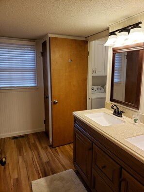 A bathroom with a wooden door, a white sink, a mirror, and a window with blinds. - Lakefront home in a Gated Community, 10 miles from Pinehurst Village (Seven Lakes)