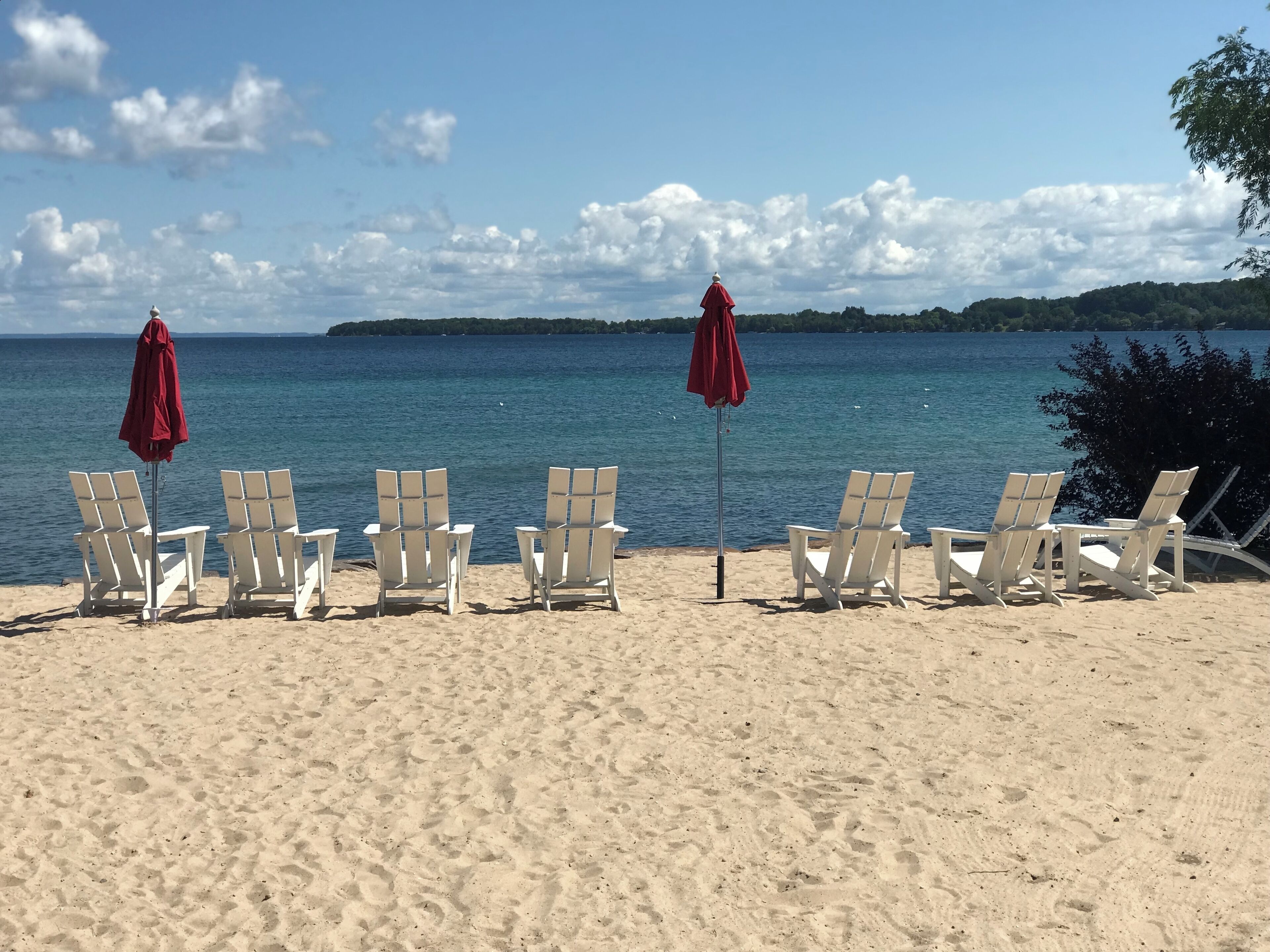Ein Strand mit weißen Liegestühlen und roten Sonnenschirmen, ein sandiger Uferstreifen und ein ruhiges Meer mit einer entfernten Küstenlinie.