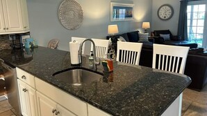 A kitchen with a granite countertop, stainless steel sink, and white cabinetry. There is a dining area with a black chair and a table, and a living area with a sofa and a lamp. - Luxury 4BR Townhouse, 2 Primary Bedrooms, east of route 1, POOL,  close to beach (Rehoboth Beach)