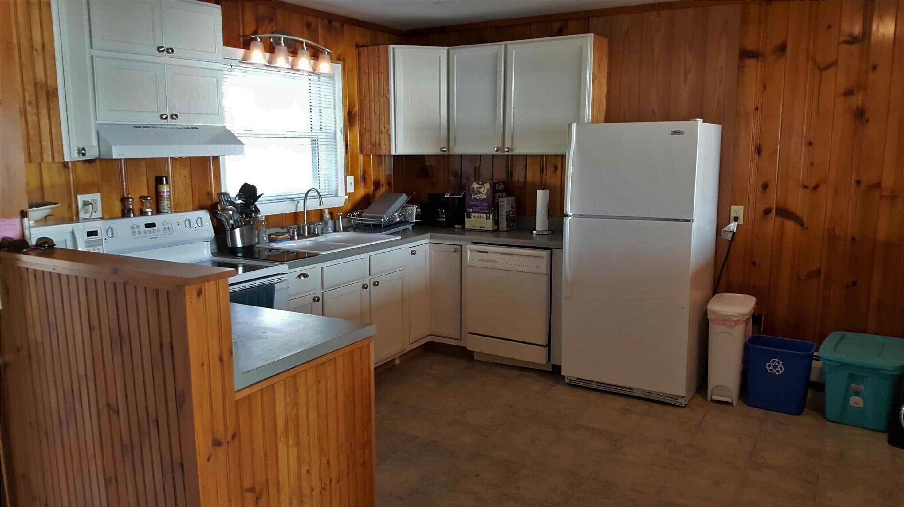 Una cocina con paneles de madera, armarios blancos, un refrigerador blanco, un lavavajillas y un fregadero con ventana arriba.