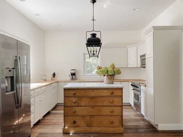 A modern kitchen with white cabinetry, a stainless steel refrigerator, a wooden island with drawers, and a hanging light fixture.