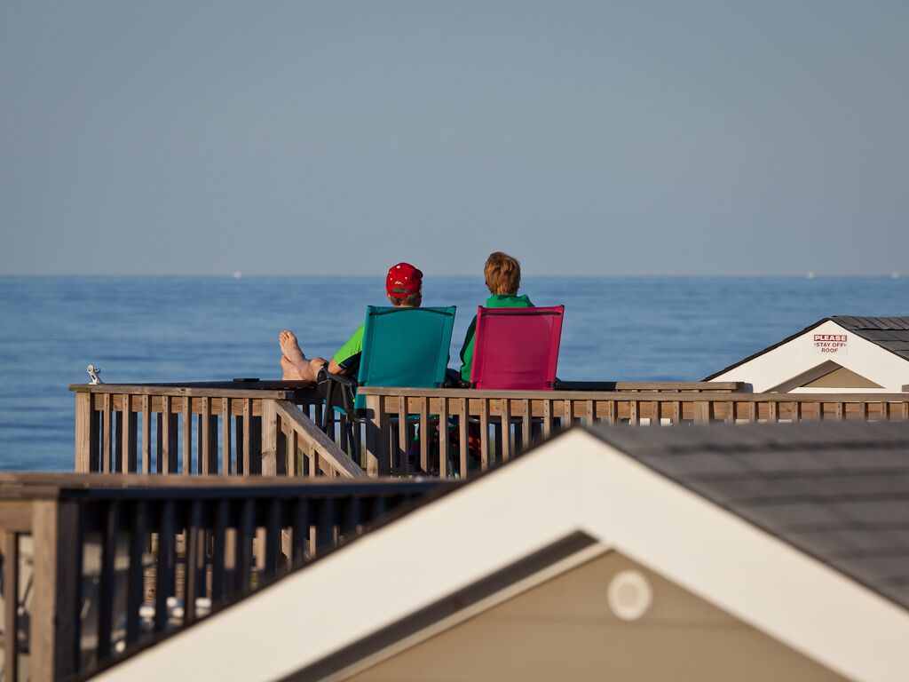 Two people sitting on a wooden deck overlooking the sea, with a building labeled "STAY OFF POOL" in the background.