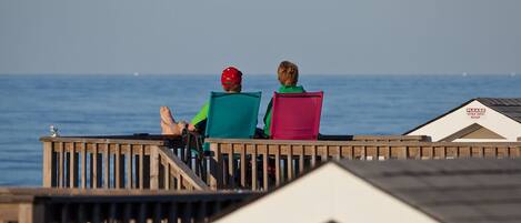 Two people sitting on a wooden deck overlooking the sea, with a building labeled "STAY OFF POOL" in the background.