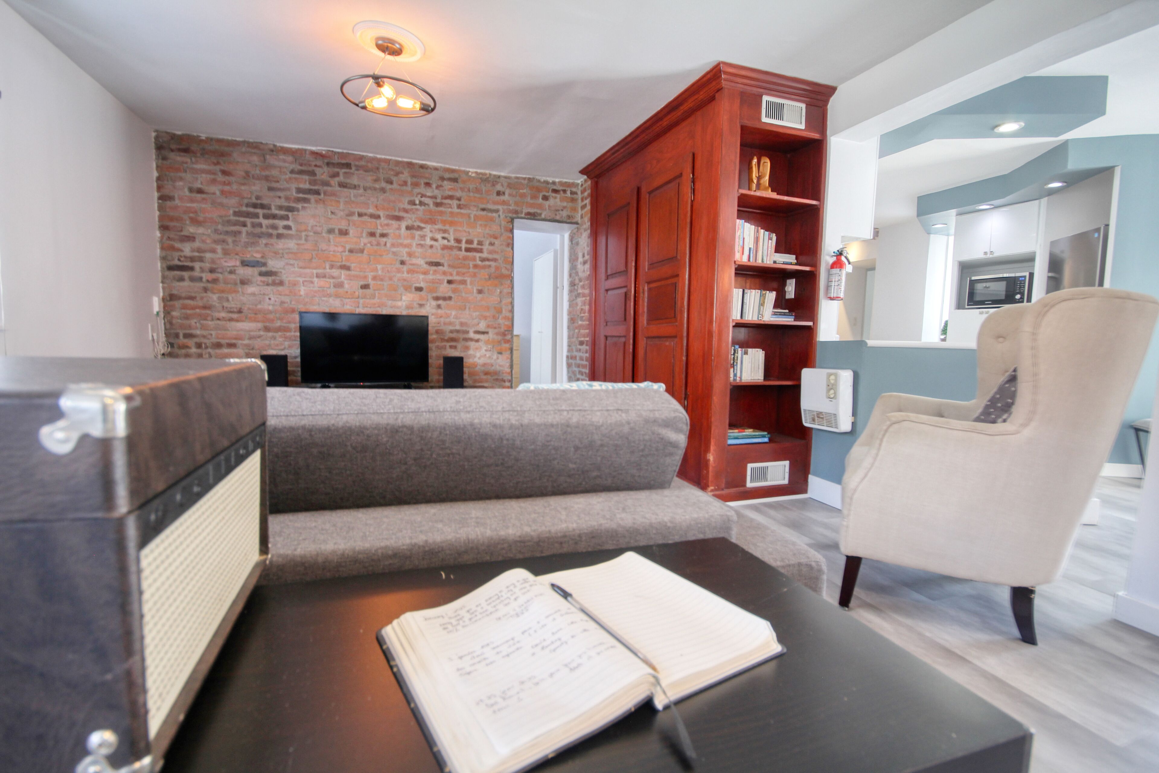 Un salon avec un mur en briques, un téléviseur à écran plat, un canapé, une bibliothèque en bois et un fauteuil beige.